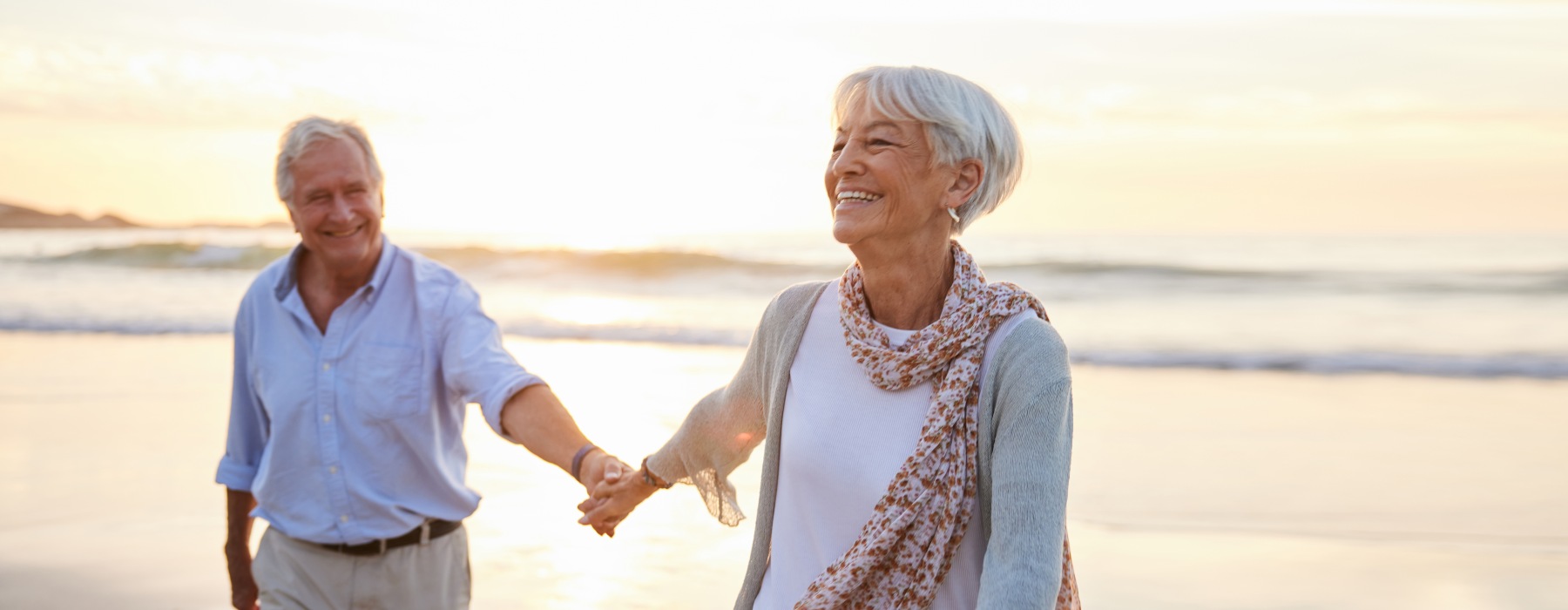 A happy senior couple smiling and holding hands while walking along the ocean shore at sunset near Dream Portofino Shores.