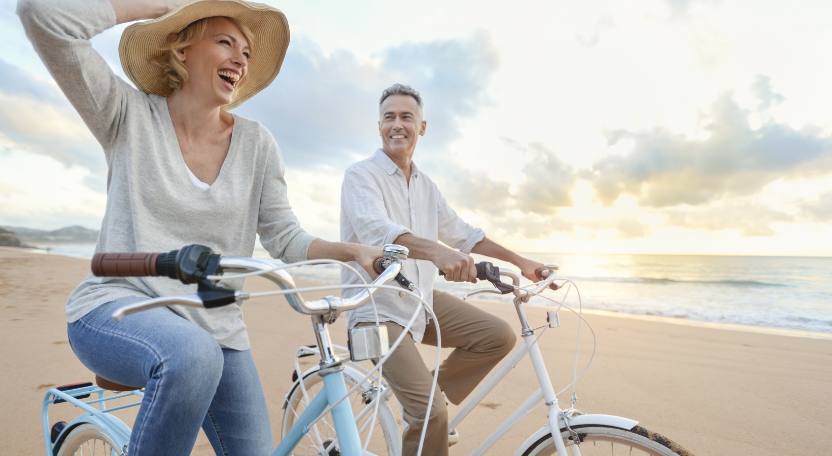 An active senior couple riding cruiser bicycles along a sandy beach at sunset near Portofino Shores.